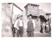 Two men standing talking in front of structure made of logs