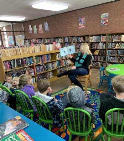Library Directory Cori Adams Reading to a group of children seated in green chairs for storytime.